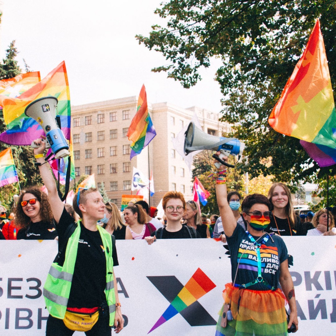 A cheerful crowd of people standing behind a colorful banner. There are LGBT+ Pride flags and hand-made posters. Two women stand in front of the banner. Each has a hand raised, with a megaphone in it.