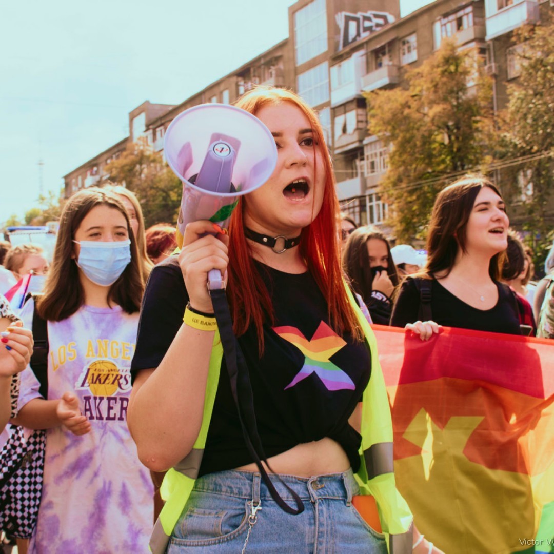 A march volunteer wearing a black t-shirt with the rainbow logo. It is a KharkivPride logo. It is a rainbow light beam going through a gray concrete rectangle.