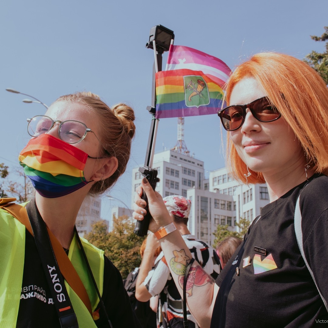 Two women are holding cameras to report from the Pride March. They are wearing branded t-shirts and rainbow face masks because of COVID restrictions. One woman is holding two flags. The first flag is the Kharkiv coat of arms on the rainbow background. The second flag is the Lesbian Pride flag with bent lines and the KharkivPride logo.