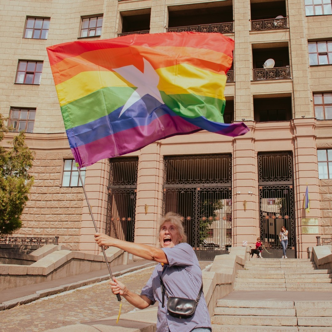 A passionate woman is waving a huge Pride flag. The flag has bent rainbow lines and a big white KharkivPride logo at the center.